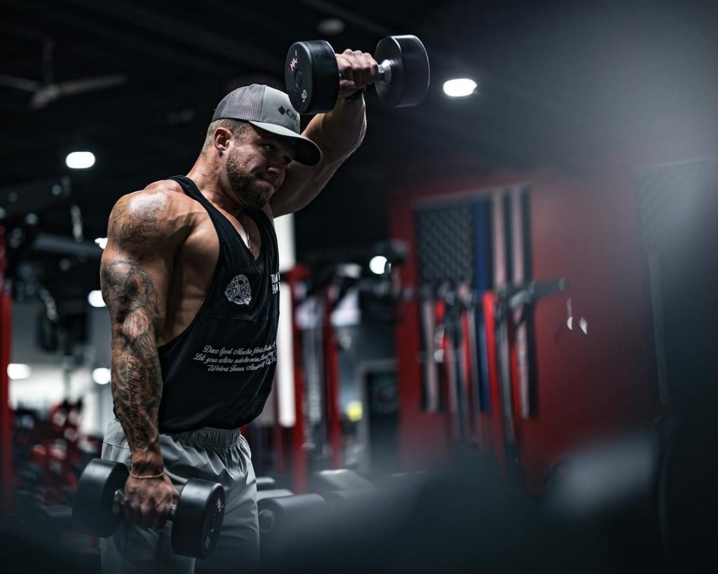 A muscular man with tattoos performs dumbbell exercises in a modern gym environment.