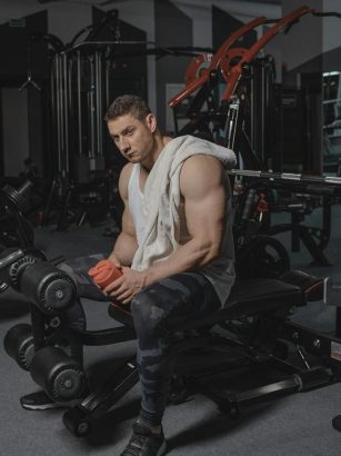 Man seated on bench in gym holding protein shaker, surrounded by equipment.