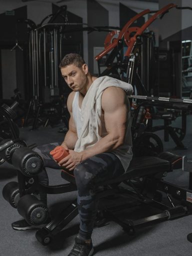 Man seated on bench in gym holding protein shaker, surrounded by equipment.